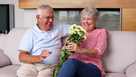 Senior-man-surprising-partner-with-flowers-on-the-couch-