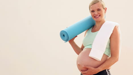 Pregnant-blonde-smiling-at-camera-holding-exercise-mat