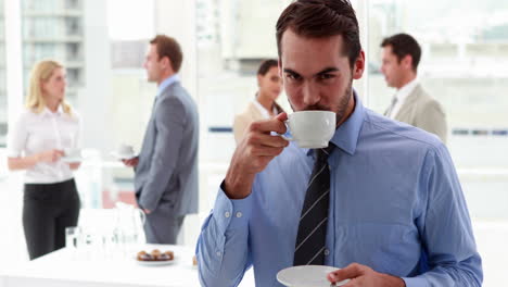 Businessman-standing-and-drinking-a-coffee-with-colleagues-behind