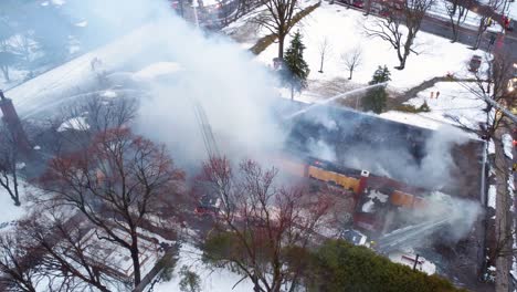 Luftaufnahme-Einer-Drohne,-Die-Nach-Einem-Brand-Auf-Einen-Schwelenden,-Verlassenen-Kindergarten-Herabsteigt,-Während-Feuerwehrleute-Weiterhin-Den-Brand-Löschen