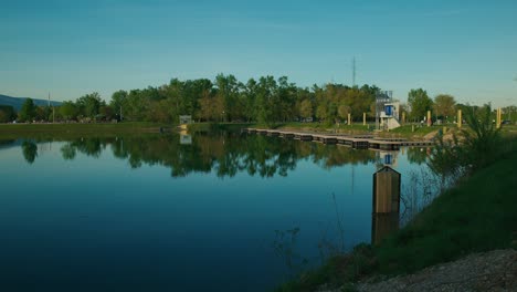 Reflejos-De-árboles-Y-Una-Estructura-Moderna-En-Un-Lago-Tranquilo-Con-Un-Cielo-Despejado-En-El-Lago-Jarun,-Zagreb