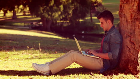 Junger-Mann-Sitzt-An-Einem-Baum-Und-Benutzt-Seinen-Laptop