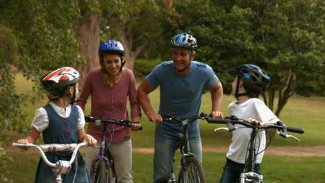 Glückliche-Familie-Auf-Ihrem-Fahrrad-Im-Park-Lächelnd-In-Die-Kamera