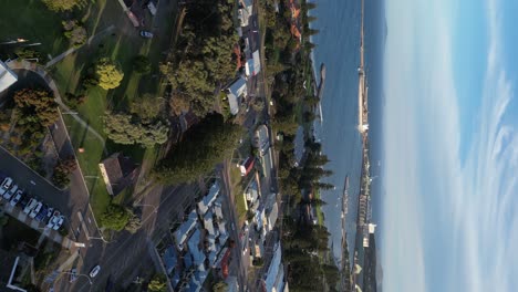 Urban-area-with-moving-cars-surrounded-by-trees-near-the-seaside-and-seaport,-aerial-dynamic-vertical-shot