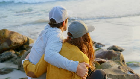 Vista-Trasera-De-Una-Joven-Pareja-Caucásica-Sentada-En-Una-Roca-Y-Mirando-El-Mar-En-La-Playa-4k