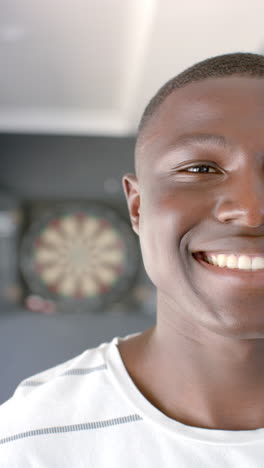 Vertical-video:-A-young-African-American-man-smiles-broadly,-dartboard-in-the-background