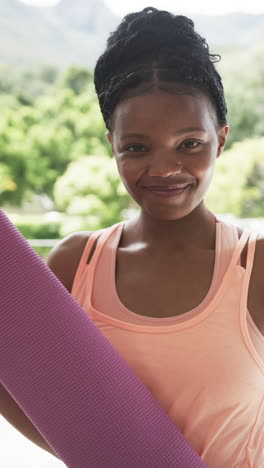 Vertical-video:-Young-African-American-woman-holding-yoga-mat,-smiling-broadly-at-home