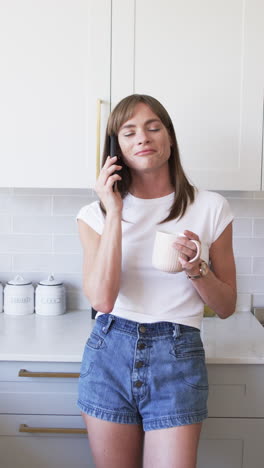 Vídeo-Vertical:-Mujer-Caucásica-Hablando-Por-Teléfono,-Sosteniendo-Una-Taza-En-Casa-En-La-Cocina