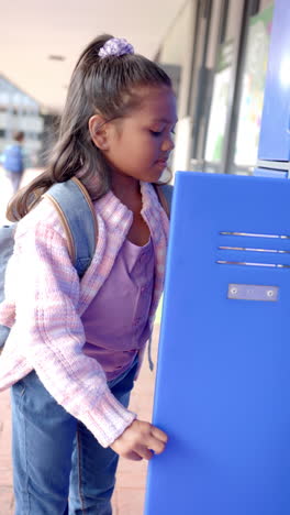 Vertical-video:-In-school,-young-biracial-girl-with-light-brown-skin-is-opening-a-blue-locker