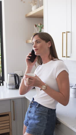 Vertical-video:-Caucasian-woman-talking-on-smartphone,-at-home-in-the-kitchen