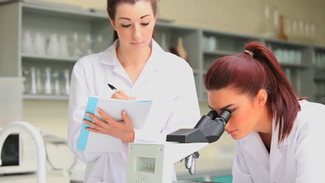 Science-student-looking-in-a-microscope-while-his-classmate-is-writing