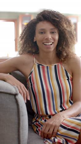 Vertical-video:-A-young-biracial-woman-with-curly-brown-hair-smiles-warmly-at-home-on-couch