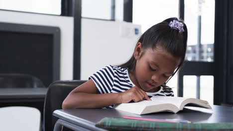In-a-school-classroom,-a-biracial-young-girl-is-focused-on-reading-a-book