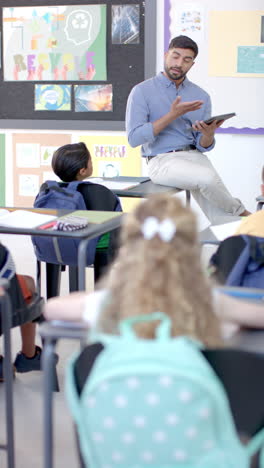 Asian-male-teacher-with-short-black-hair-sits-on-desk,-hand-raised-in-vertical-video