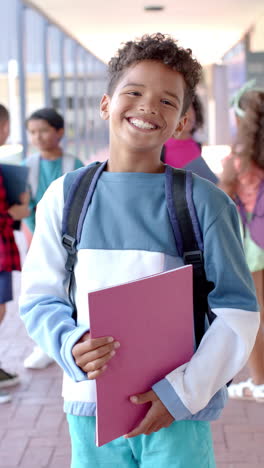 Vertical-video:-In-school,-young-boy-holding-books-and-wearing-a-backpack-is-smiling