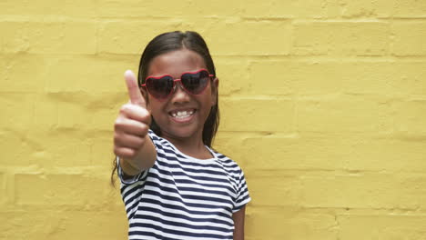 A-young-biracial-girl-stands-before-a-yellow-background,-giving-a-thumbs-up