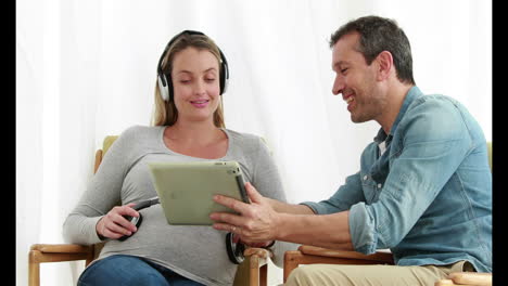 Un-Hombre-Usando-Una-Tableta-Mientras-Una-Mujer-Embarazada-Escucha-Música-Con-Auriculares