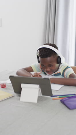 Vertical-video:-African-American-boy-wearing-headphones,-looking-at-tablet-and-doing-homework