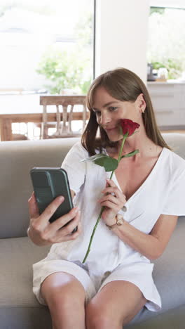 Vertical-video:-Caucasian-woman-holding-red-rose,-smiling-at-phone-at-home