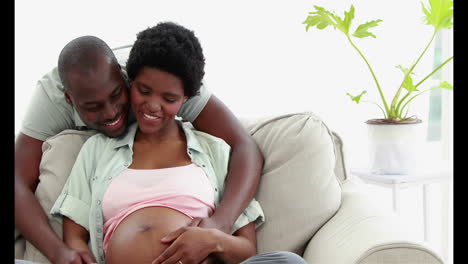Smiling-pregnant-woman-sitting-on-the-couch-while-embracing-her-husband