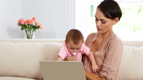 Linda-Madre-Sosteniendo-A-Su-Bebé-Y-Usando-Una-Computadora-Portátil