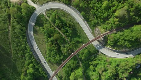 Static-aerial-overhead-footage-of-multiple-cars-driving-on-a-winding-road-intersected-by-an-old-railway-bridge-in-the-middle-of-a-forest