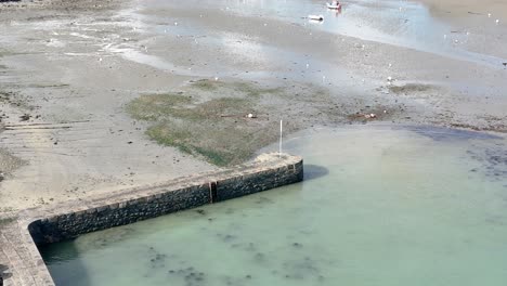 Niedrig-Fallende-Und-Kreisende-Drohnenaufnahme-über-Dem-Hafen-Von-Bordeaux,-Guernsey-Bei-Ebbe-Mit-Hafenmauer,-Anlegestelle,-Auf-Sand-Trocknenden-Booten,-Strand-Und-Hütten-Im-Hintergrund-An-Einem-Hellen-Tag