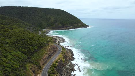 4K-Video-of-a-lone-camper-van-driving-through-the-beautiful-Great-Ocean-Road-with-the-green,-lush-nature-on-one-side-of-the-road-and-the-blue-ocean-waves-crashing-on-the-other-side