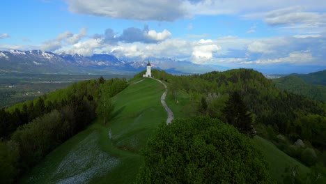 Lush-green-hill-with-a-winding-path-leading-to-a-solitary-white-church,-set-against-a-backdrop-of-majestic-mountains-and-a-cloudy-sky