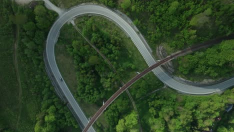 Static-aerial-overhead-footage-of-one-car-driving-on-a-winding-road-intersected-by-an-old-railway-bridge-in-the-middle-of-a-forest