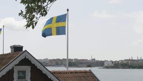 Bandera-Sueca-Junto-Al-Mar-De-Karlskrona-Con-Fondo-De-Paisaje-Urbano,-Blekinge,-Suecia