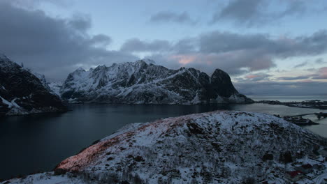 Stunning-drone-flight-over-Reine,-Lofoten-Islands,-Norway,-featuring-snowy-mountains,-the-ocean,-and-a-cloudy-sky