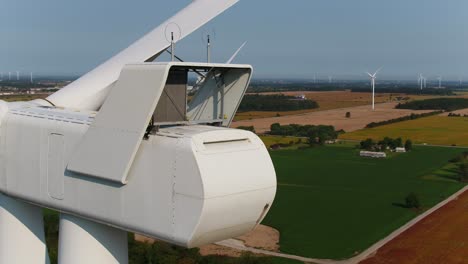 Windpark-Mit-Nahaufnahme-Der-Gondel-Zur-Inspektion-Mithilfe-Einer-Drohne,-Die-Die-Maschine-Umkreist,-Vor-Ackerfeldern-Im-Hintergrund