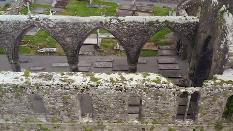 High-stone-arches-of-crumbling-ruins-in-Claregalway-Friary-covered-with-moss,-aerial-high-angle-pan