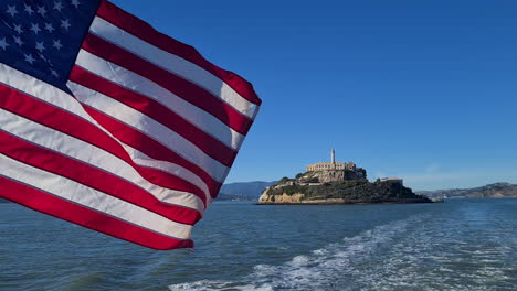 Ondear-La-Bandera-Americana-En-El-Ferry-Con-La-Isla-De-Alcatraz-Y-La-Prisión-En-El-Fondo,-California,-EE.UU.