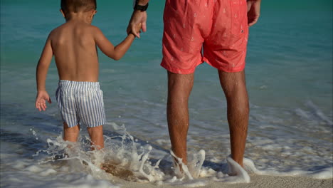 Slow-motion-of-a-small-mexican-latin-boy-holding-his-father's-hand-standing-at-the-beach-waiting-for-the-waves-to-hit-their-legs