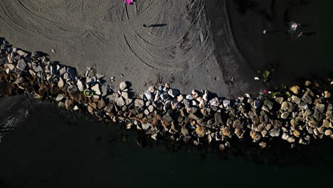 Rock-breakwater-and-sandy-beach,-aerial-top-down-ascend-view