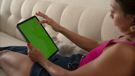 Slow-motion-of-a-mexican-latin-woman-with-a-pink-top-laying-on-her-couch-scrolling-browsing-her-tablet-with-her-index-finger-going-upwards