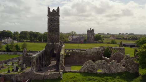 Claregalway-Friary-view-from-behind-with-castle-in-distance,-drone-dolly-over-cemetery