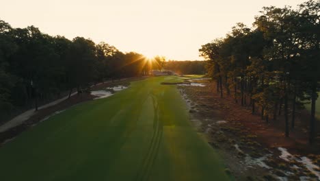 aerial-drone-shot-down-shot-moving-down-the-fairway-of-a-golf-course-as-the-rising-sun-peaks-over-the-trees