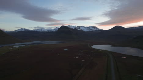 Beautiful-drone-flight-over-rural-Iceland-at-sunset,-showcasing-snowy-mountains,-green-fields,-lakes,-and-a-stunning-sunset-sky