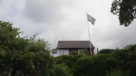 Bandera-Sueca-Ondeando-En-Lo-Alto-De-Un-Asta-De-Bandera-En-Una-Cabaña-De-Verano,-Vista-De-ángulo-Bajo