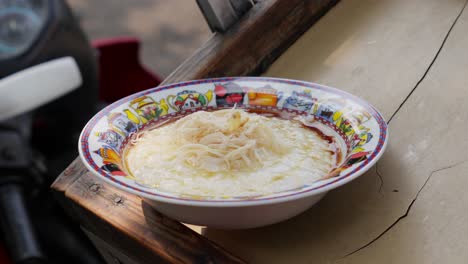 A-tongs-placing-shredded-chicken-on-rice-porridge-in-a-colorful-bowl-on-a-wooden-surface