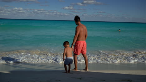 Zeitlupe-Eines-Mexikanisch-lateinamerikanischen-Vaters-Und-Seines-Sohnes,-Die-Händchen-Haltend-Am-Strand-Stehen-Und-An-Einem-Sonnigen-Nachmittag-In-Cancun-Auf-Das-Meer-Blicken