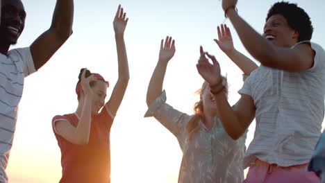 Diverse-group-of-friends-celebrate,-raising-hands-against-a-sunset-sky