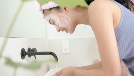 Young-biracial-woman-rinses-her-face-with-water-at-a-sink,-her-skin-covered-in-soap