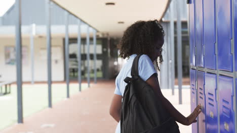 Chica-Birracial-Con-Pelo-Rizado-En-Los-Casilleros-De-La-Escuela,-Sosteniendo-Un-Libro-Rojo