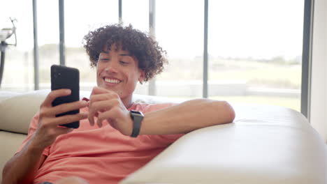 A-young-biracial-man-is-engrossed-in-his-smartphone-while-relaxing-on-a-couch-at-home