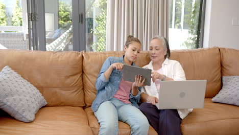 Abuela-Asiática-Y-Nieta-Birracial-Sentadas-En-El-Sofá,-Usando-Dispositivos