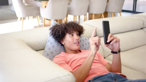 A-young-biracial-man-is-lounging-on-sofa,-smartphone-in-hand